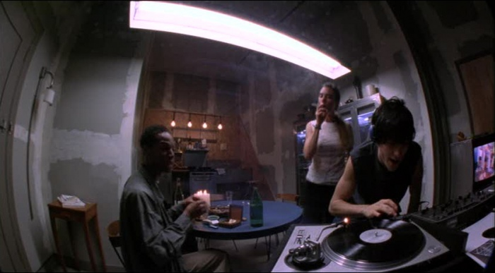 Fisheye lens shot from Requiem for a Dream of a room with three people, one sitting at a table, one standing and smoking, and one DJing.